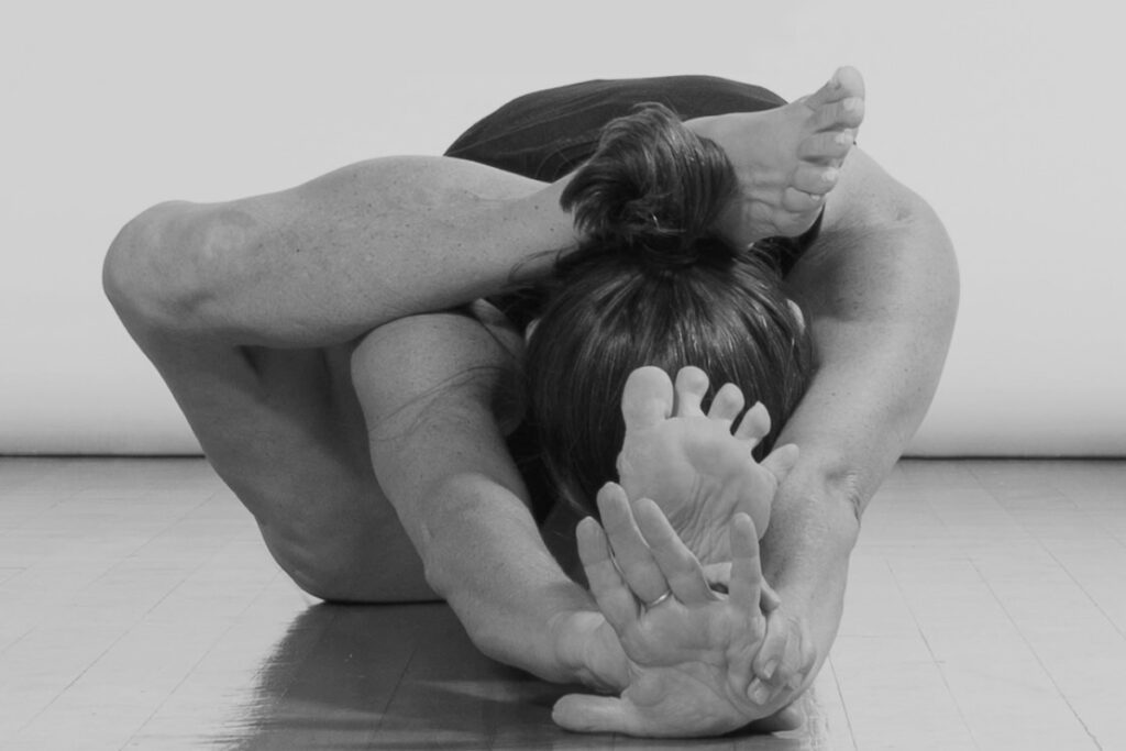 Schwarzweißfoto von Yogalehrerin Marina Jung, sie übt Vorbeuge, das rechte bein ruht mit dem Knöchel auf ihrem Nacken