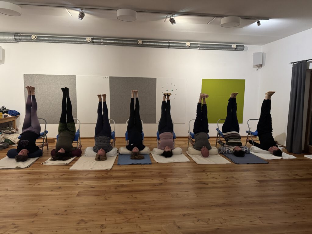 8 Iyengar-Yoga-Schülerinnen und Schüler üben Viparita Karani im Stuhl. Sie befinden sich in einer Reihe nebeneinander in einem Seminarraum