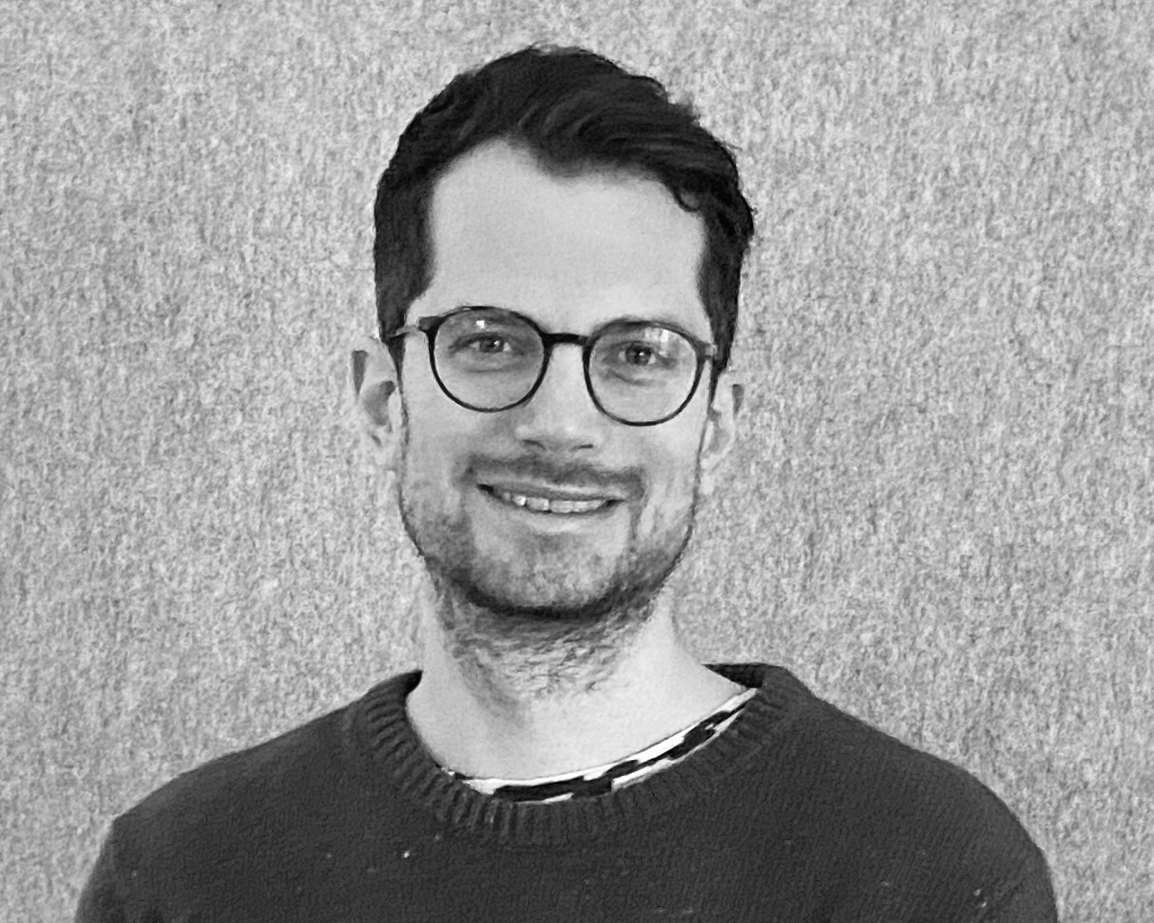 Portrait eines dunkelhaarigen jungen Mannes mit Brille und dunklem Pullover, grauer Hintergrund