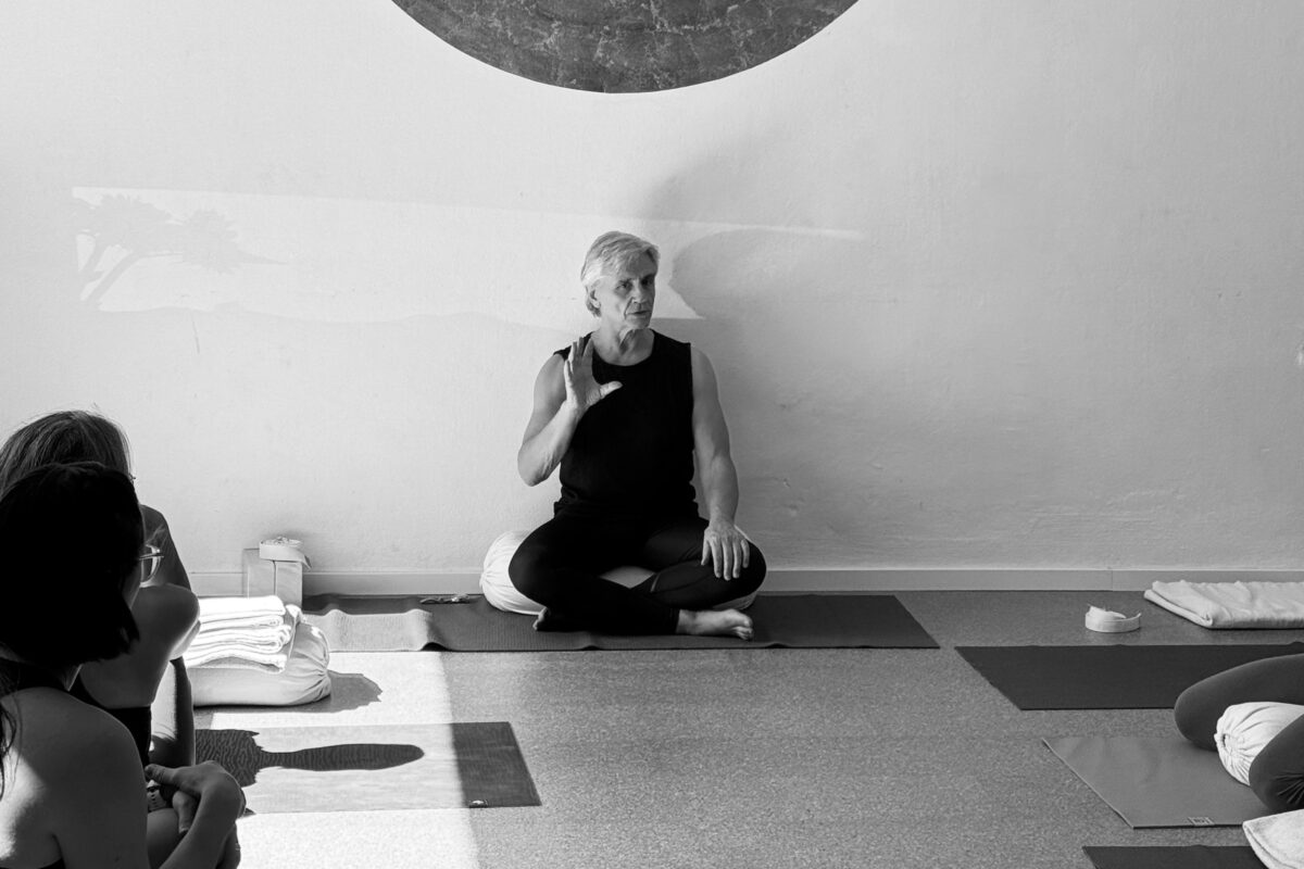 Glenn Ceresoli sitzt an eine Wand gelehnt im Schneidersitz auf einer Kissenrolle und spricht, links und rechts sieht man Schüler*innen, die ihm zuhören