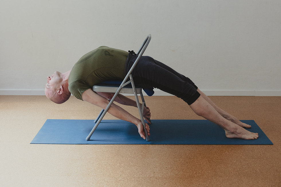 Mann in T-Shirt und Turnhose übt eine Rückbeuge mit dem Yoga-Stuhl