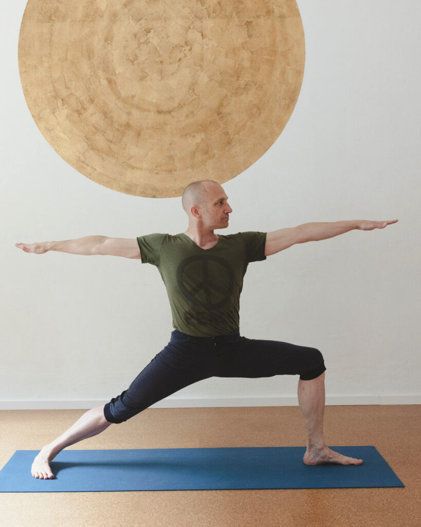 Tobias Fehrenbach steht im Krieger 2, im Yogashala, vor der goldenen Sonne an der Wand